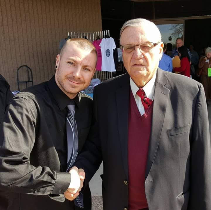 Chance Trahan (left, black shirt and tie) smiling while shaking hands with Joe Arpaio (right, suit and red sweater vest) outdoors